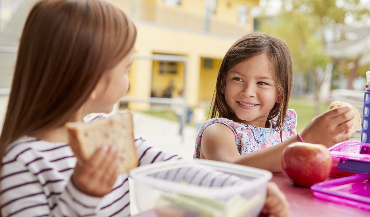 Two small girls are sitting at a table with lunchboxes filled healthy foods.