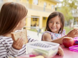 Two small girls are sitting at a table with lunchboxes filled healthy foods.