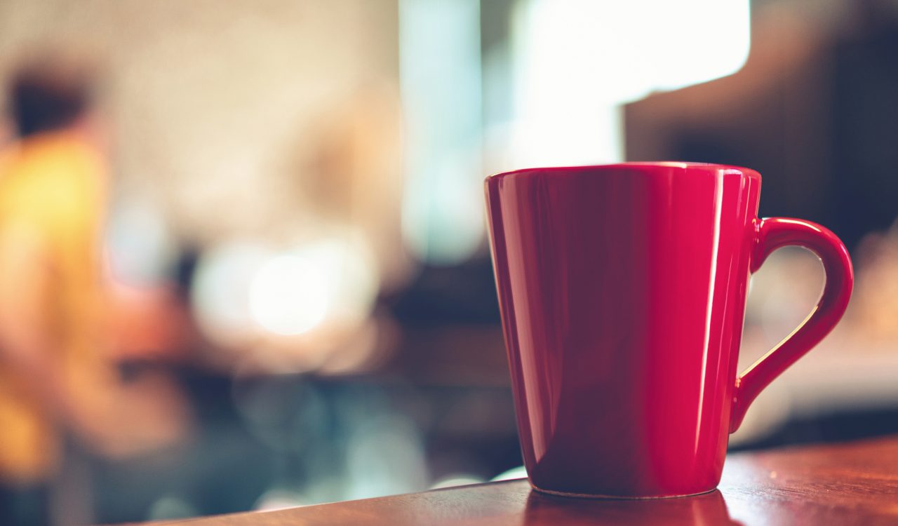 A red mug on a desk with an out-of-focus room in the background.
