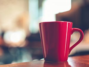 A red mug on a desk with an out-of-focus room in the background.