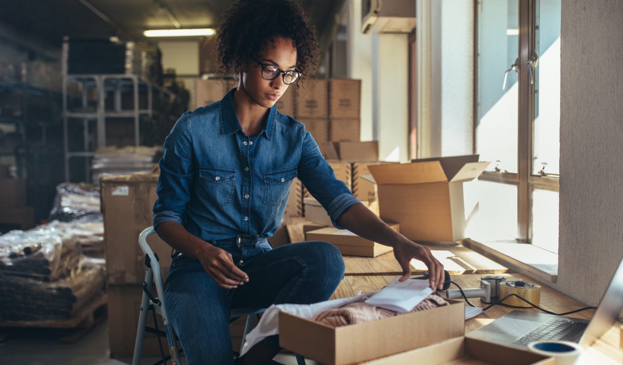 A woman sits on a stepladder leaning forward and looking down at a package she’s putting together on a table in front of her, with boxes and shelves in the background.