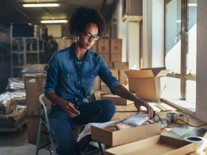 A woman sits on a stepladder leaning forward and looking down at a package she’s putting together on a table in front of her, with boxes and shelves in the background.