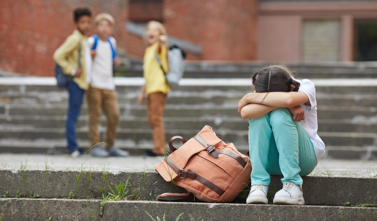 In the foreground, a girl sits on a step, hunched over, her arms covering her bent head, while three blurred-out children in the background look at her, with one of the children pointing at her.