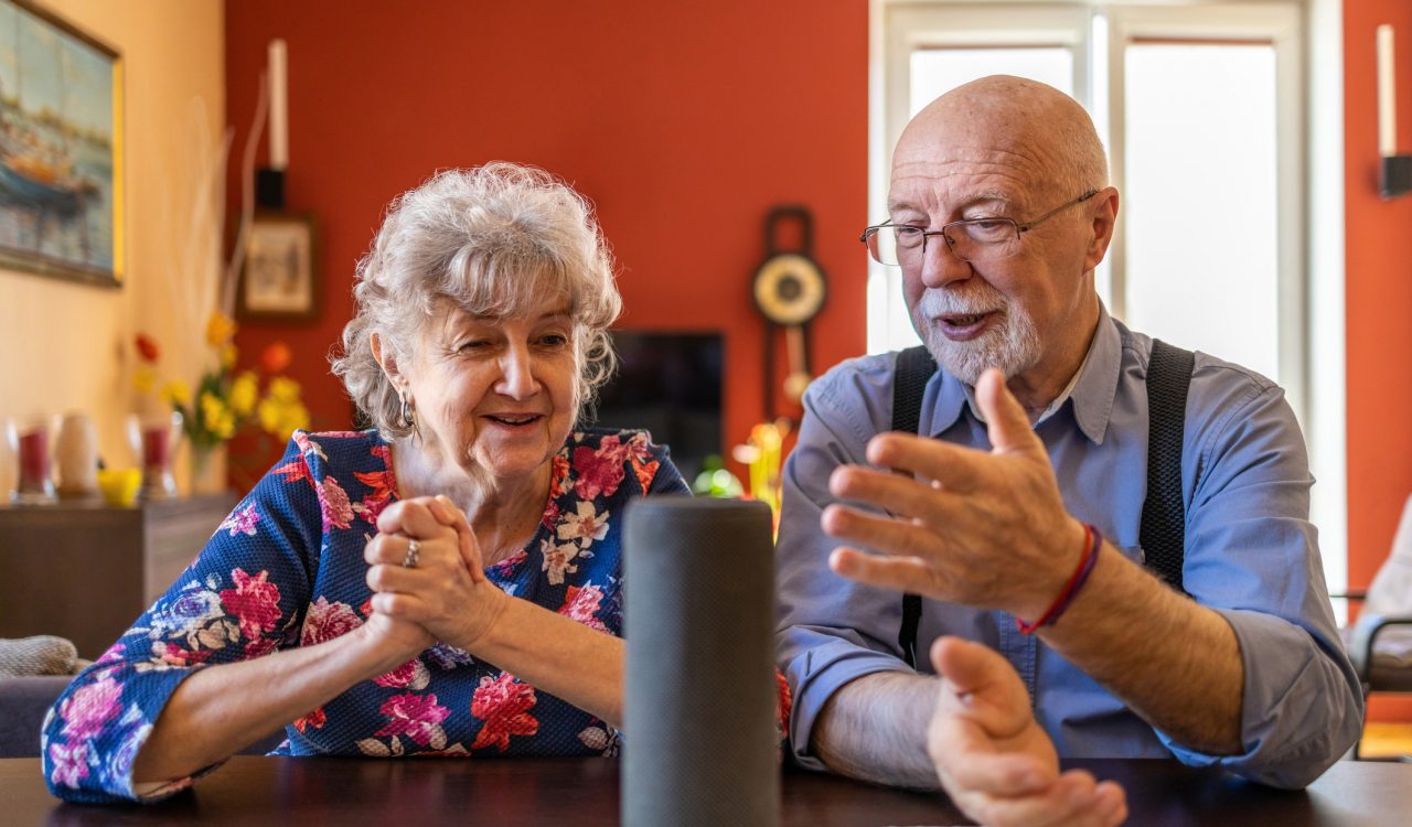 An older woman (left) and an older man (right) sit at a table smiling and looking at a virtual assistant device placed on a cloth on the table.