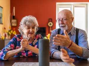 An older woman (left) and an older man (right) sit at a table smiling and looking at a virtual assistant device placed on a cloth on the table.