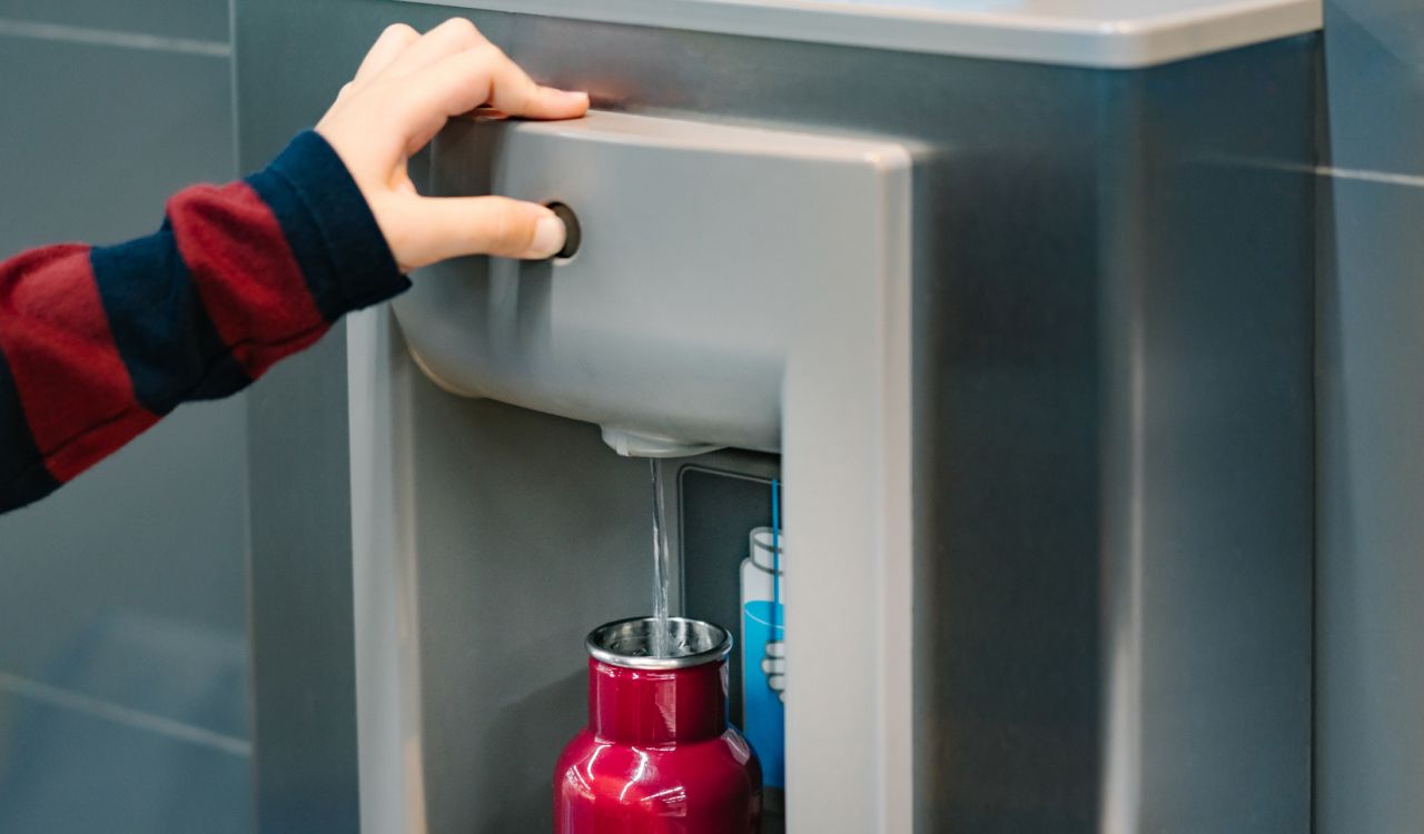 Close-up view of a hand pressing a button on a water station while water runs into a metal bottle.