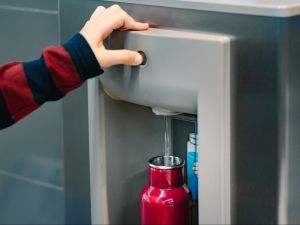 Close-up view of a hand pressing a button on a water station while water runs into a metal bottle.