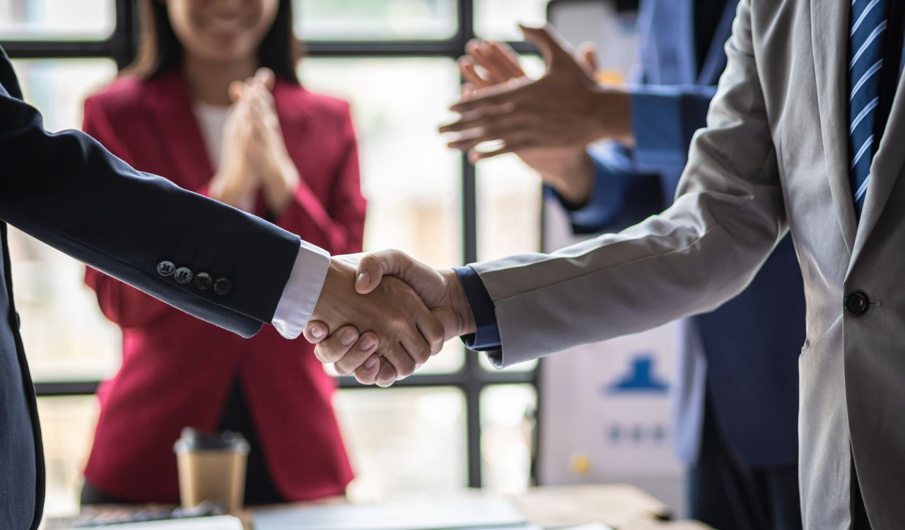 Close-up of a handshake between two people in business suits with more people in business suits applauding in the background.