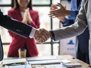 Close-up of a handshake between two people in business suits with more people in business suits applauding in the background.