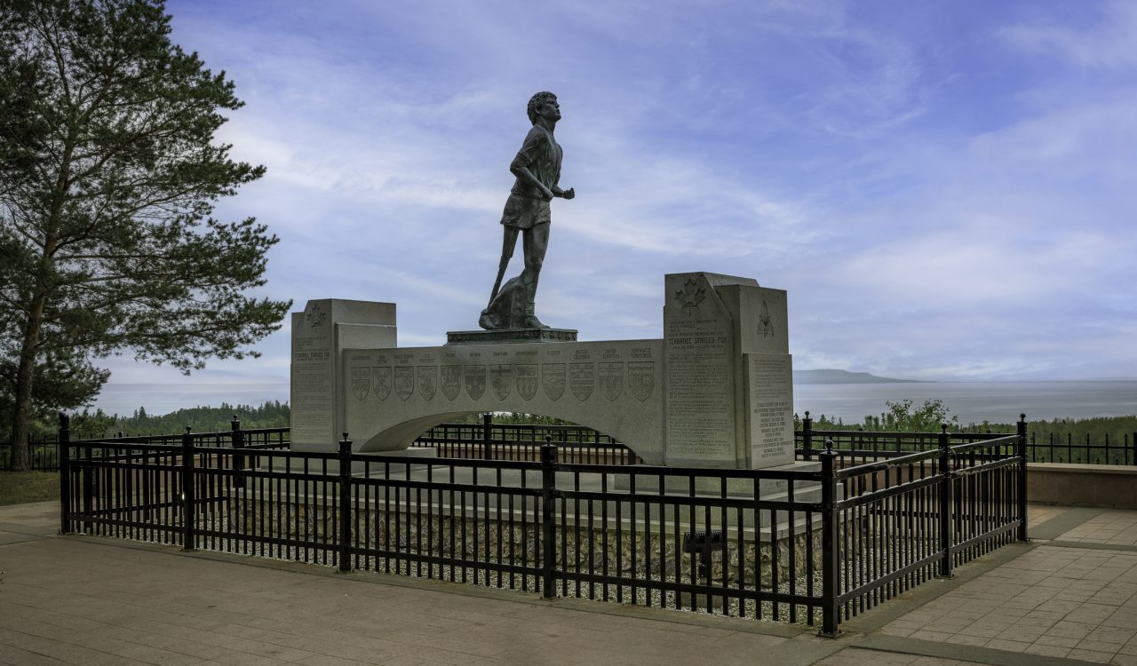 Terry Fox memorial in Thunder Bay, ON includes a large statue of Terry Fox elevated on a fenced plinth, with blue sky, trees and Lake Superior in the background.