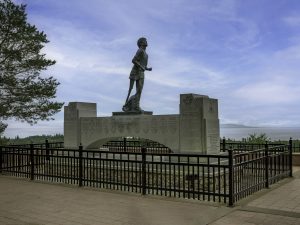 Terry Fox memorial in Thunder Bay, ON includes a large statue of Terry Fox elevated on a fenced plinth, with blue sky, trees and Lake Superior in the background.