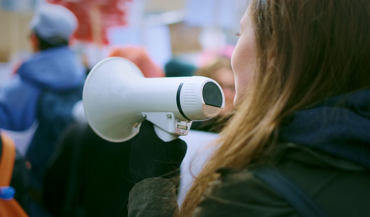 A young woman, seen from the side, speaks into a microphone in a crowd.