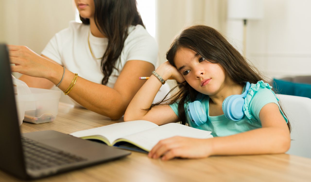 A distracted child stares at a laptop screen with a book open on the table in front of them beside their mother.