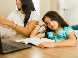 A distracted child stares at a laptop screen with a book open on the table in front of them beside their mother.