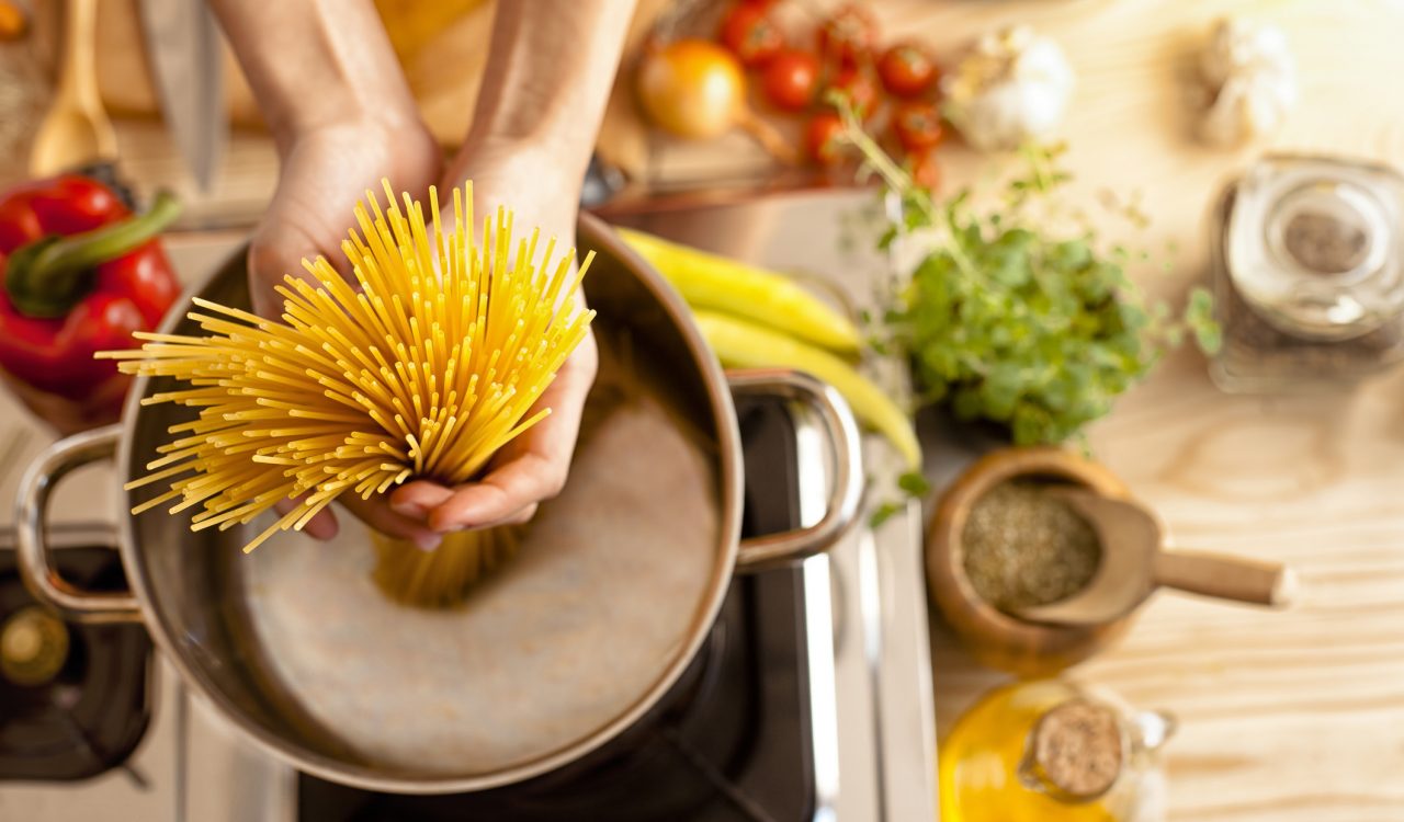 Close-up of hands adding spaghetti to a pot of boiling water with produce, herbs and spices on the counter beside it.