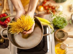 Close-up of hands adding spaghetti to a pot of boiling water with produce, herbs and spices on the counter beside it.