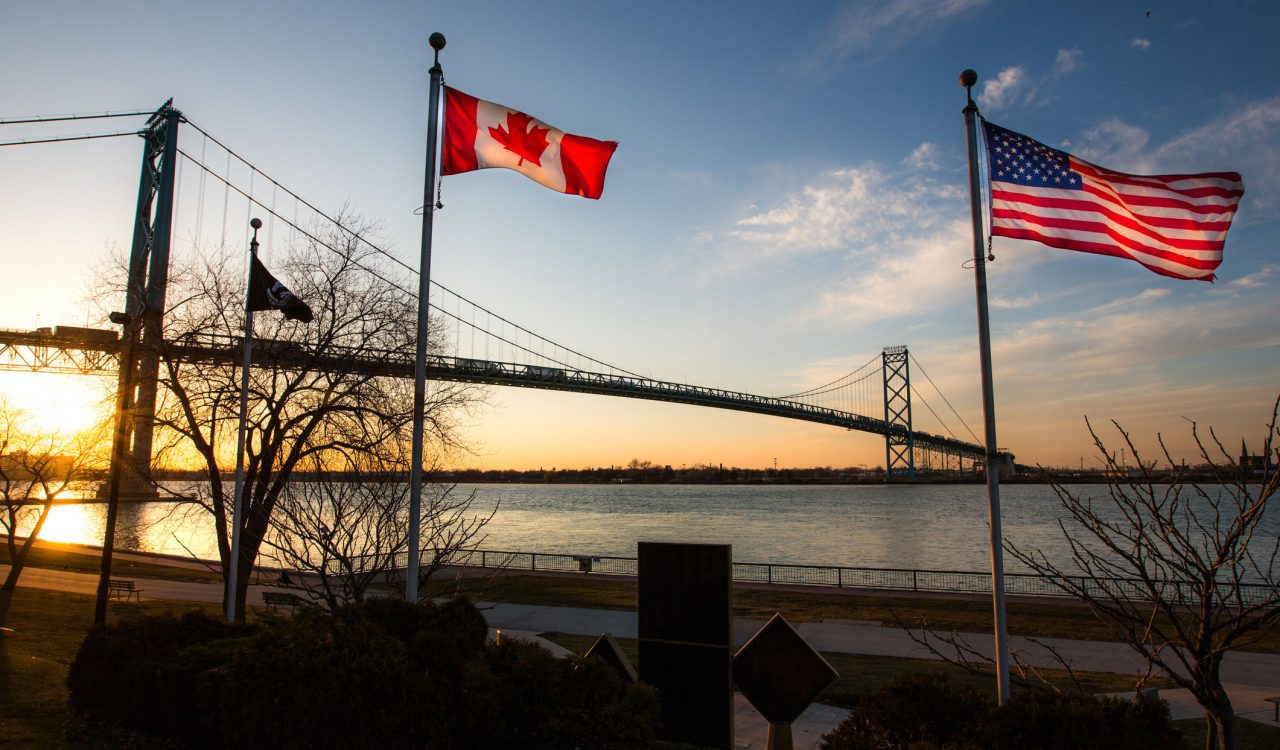 The Canadian and American flags fly from flagpoles with a bridge and sunset sky behind them.