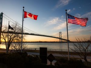 The Canadian and American flags fly from flagpoles with a bridge and sunset sky behind them.