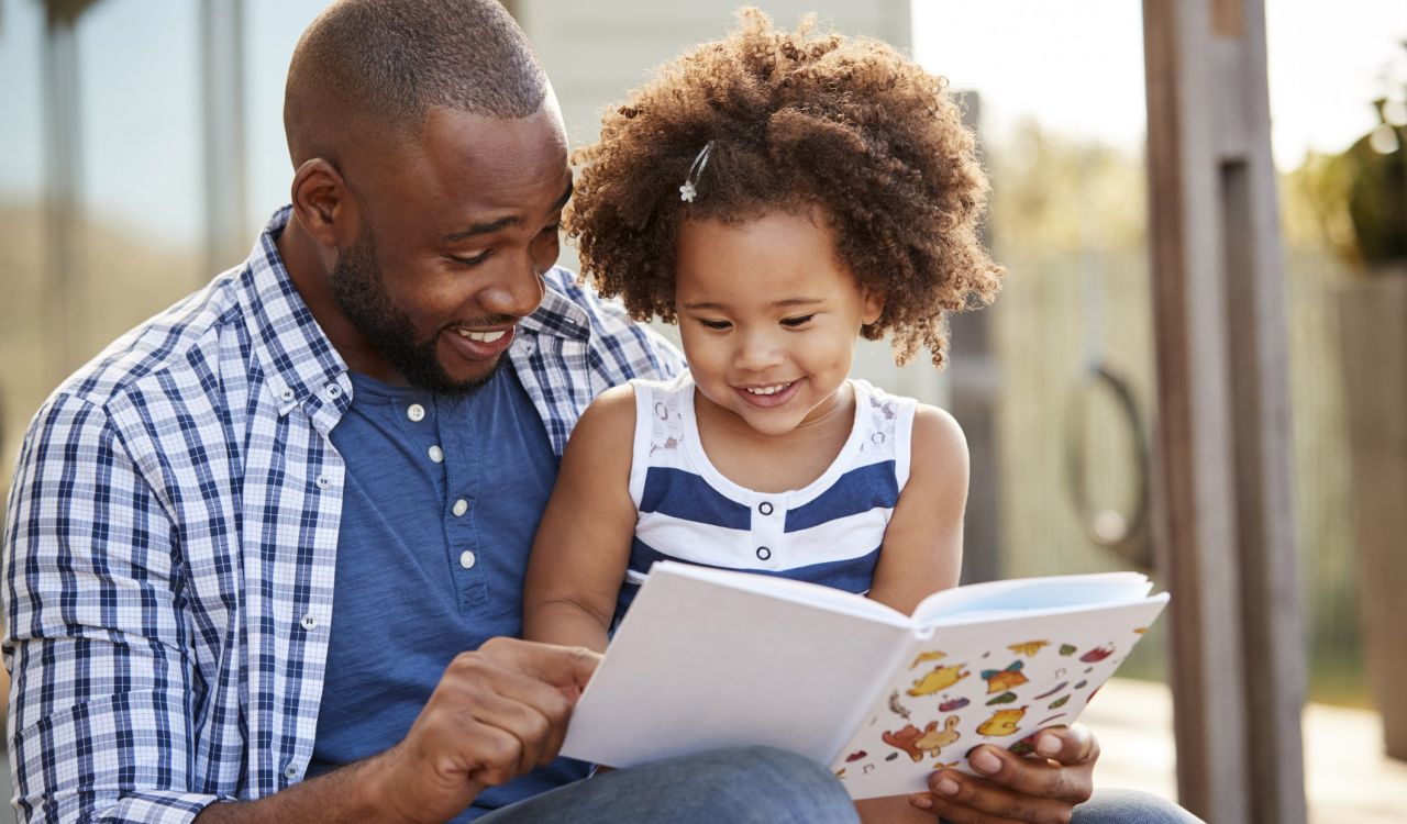 A smiling small girl sits on her smiling father’s lap and looks down at a book he’s gripping with one hand and pointing at with his other hand.