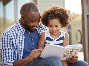 A smiling small girl sits on her smiling father’s lap and looks down at a book he’s gripping with one hand and pointing at with his other hand.