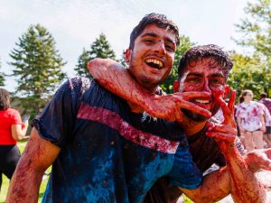 Two men laugh as they are dripping in grapes and splatter while outdoors at Brock University’s campus.