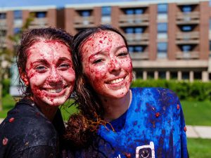 Two women smile while covered in grapes with Brock University’s Decew Residence serving as the backdrop.