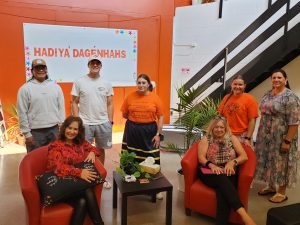 Seven people, two seated, come together in a space with a bright orange wall that reads: Hadiya’dagénhahs.