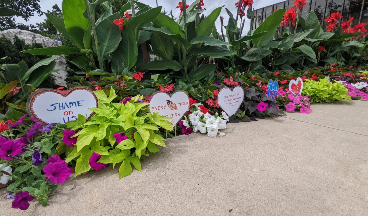 A row of paper hearts with text and images painted on them line a garden as a memorial of children lost to the residential school system.