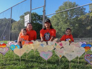 Two adults and two children, all in orange shirts, sit behind a display of hearts staked into the ground. The hearts have messages related to truth and reconciliation written on them.