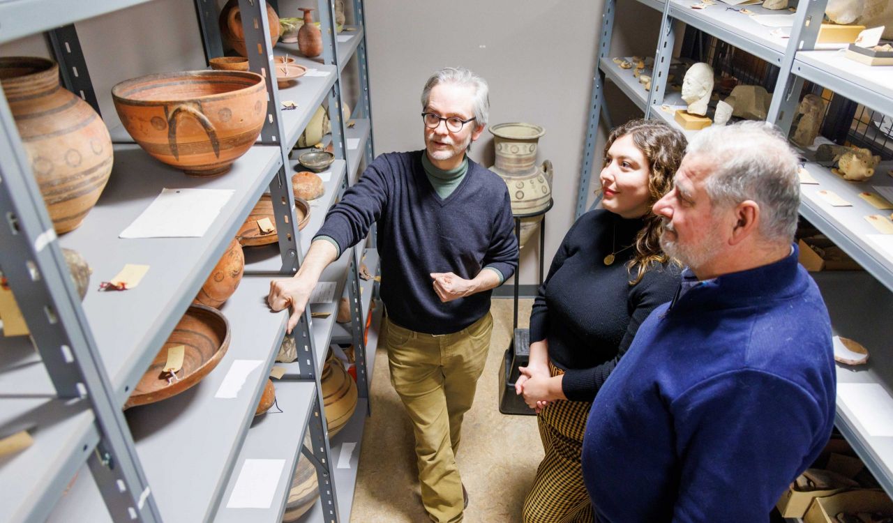 Three people stand in room looking at shelves of ancient pottery.