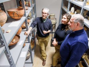 Three people stand in room looking at shelves of ancient pottery.