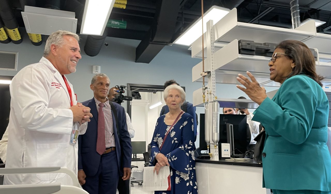 Three people in business attire and a man in a lab coat converse in a lab.