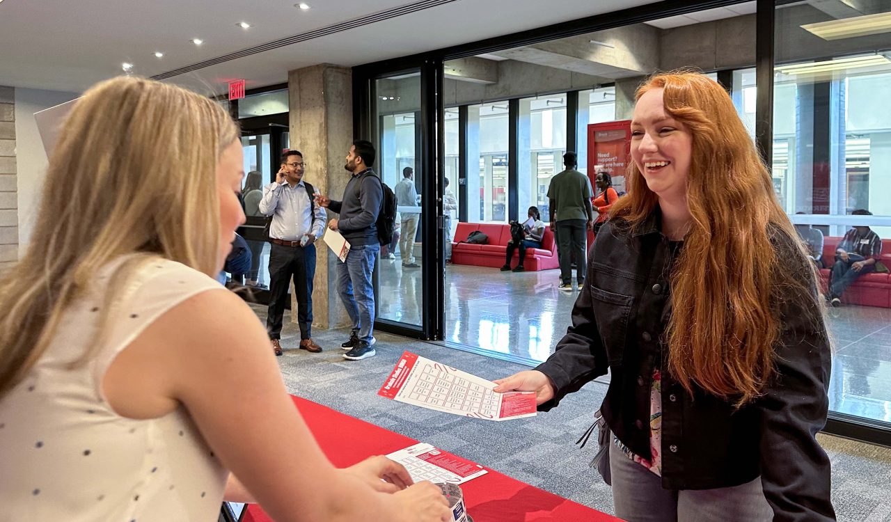 A university student receives a piece of paper from a university employee at a table in a sunny hallway.