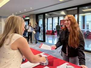 A university student receives a piece of paper from a university employee at a table in a sunny hallway.