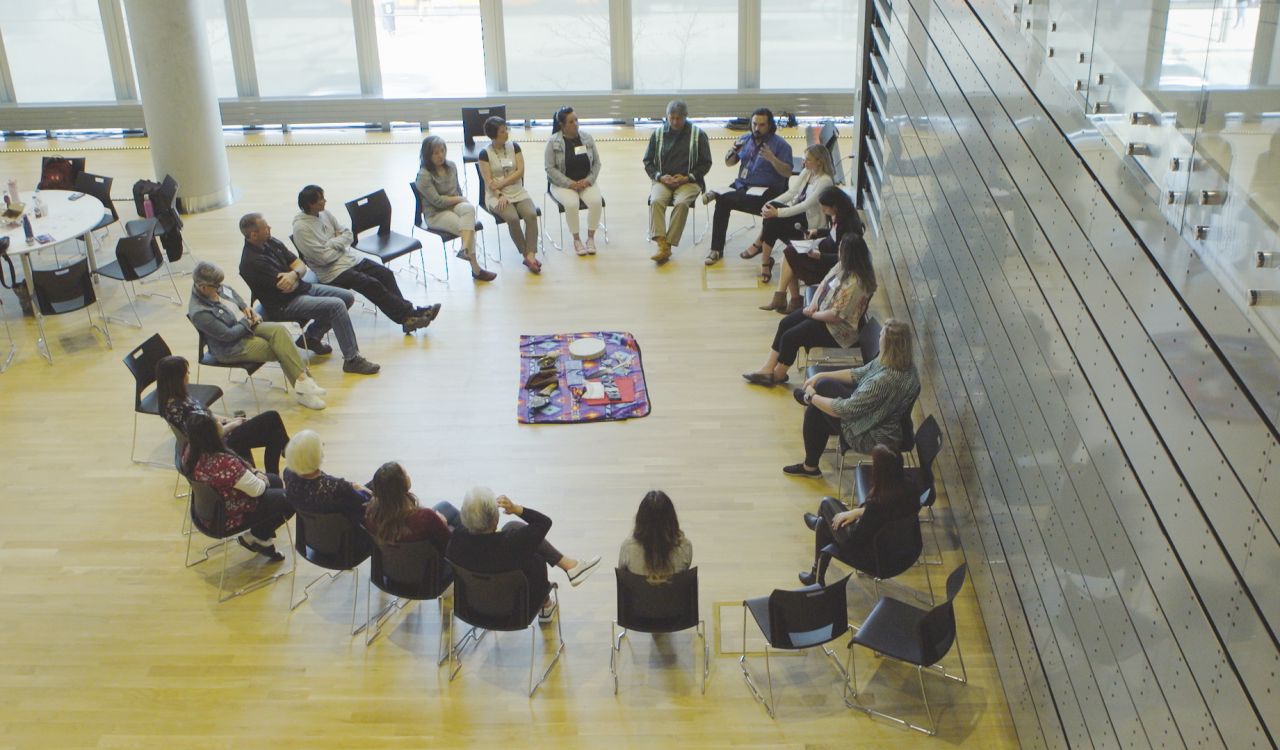 An aerial view of people sitting in a circle, with a cloth in the middle upon which lies a drum and other objects.