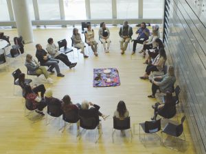 An aerial view of people sitting in a circle, with a cloth in the middle upon which lies a drum and other objects.