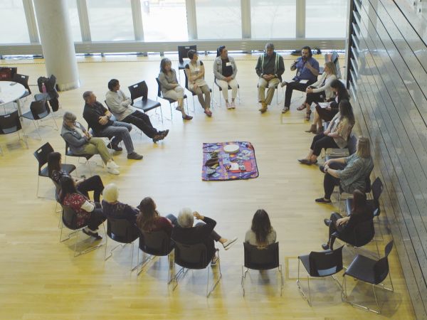 An aerial view of people sitting in a circle, with a cloth in the middle upon which lies a drum and other objects.
