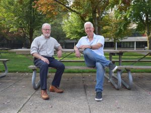 Russell Johnston and Michael Ripmeester seated on a picnic table in Jubilee Court on Brock’s main campus with trees showing a hint of fall colours in the background