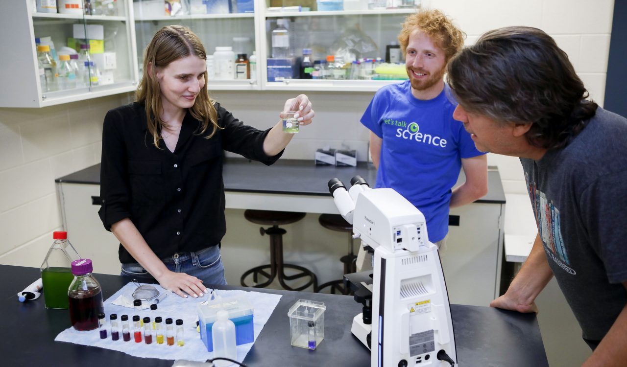 Three people stand in a science laboratory looking at equipment and materials placed on a table, such as a beaker of algae, a microscope, pipettes and colourful substances.