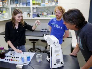 Three people stand in a science laboratory looking at equipment and materials placed on a table, such as a beaker of algae, a microscope, pipettes and colourful substances.