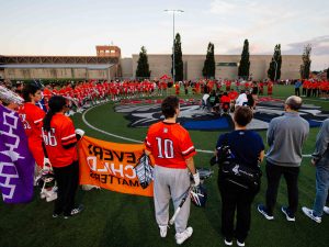 A crowd of people, including lacrosse players and spectators, stand in a large circle on a sports field with drummers in the centre.