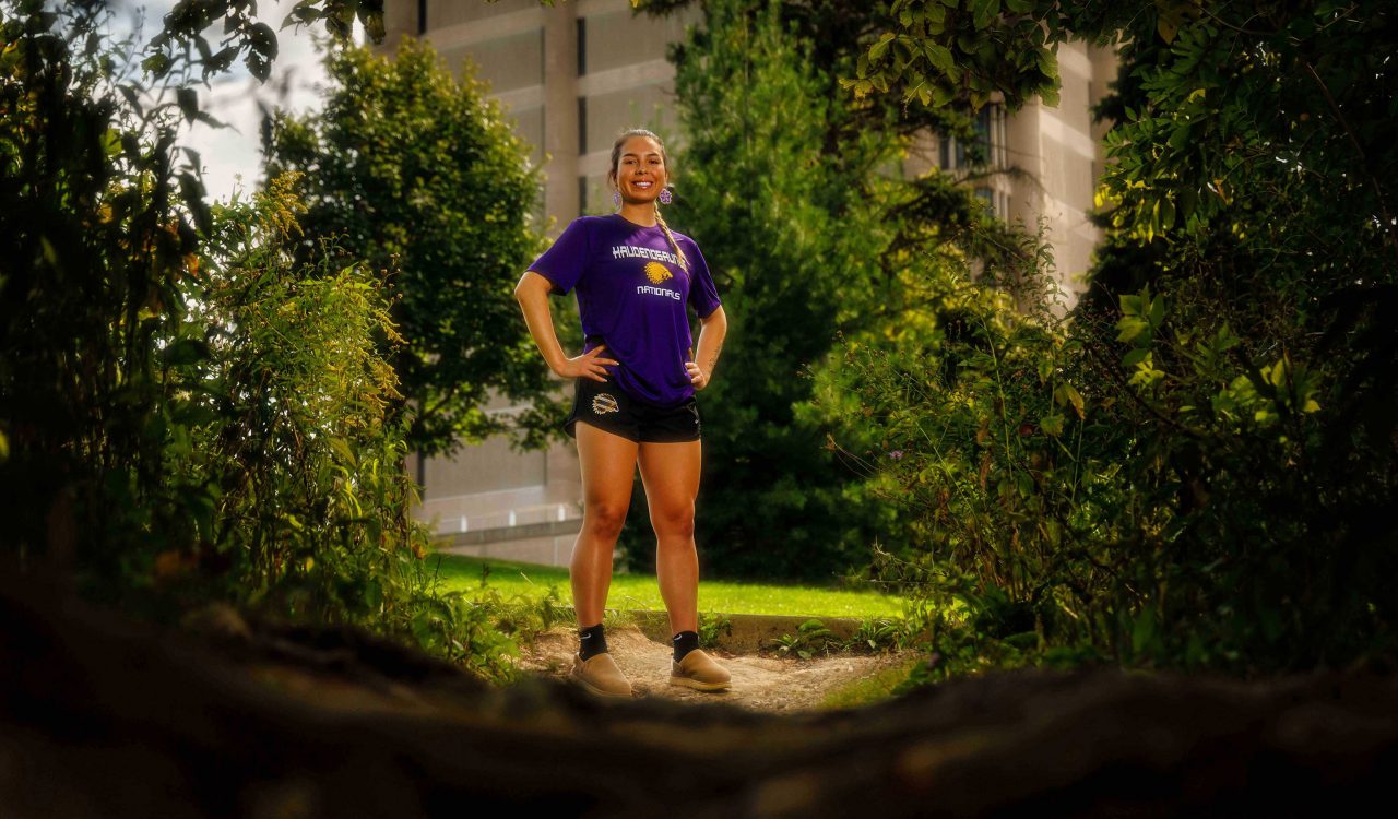 A woman stands at a trailhead framed by a forest with Brock University in the background while wearing purple lacrosse sportswear.