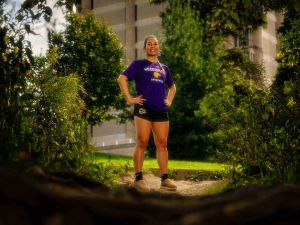 A woman stands at a trailhead framed by a forest with Brock University in the background while wearing purple lacrosse sportswear.