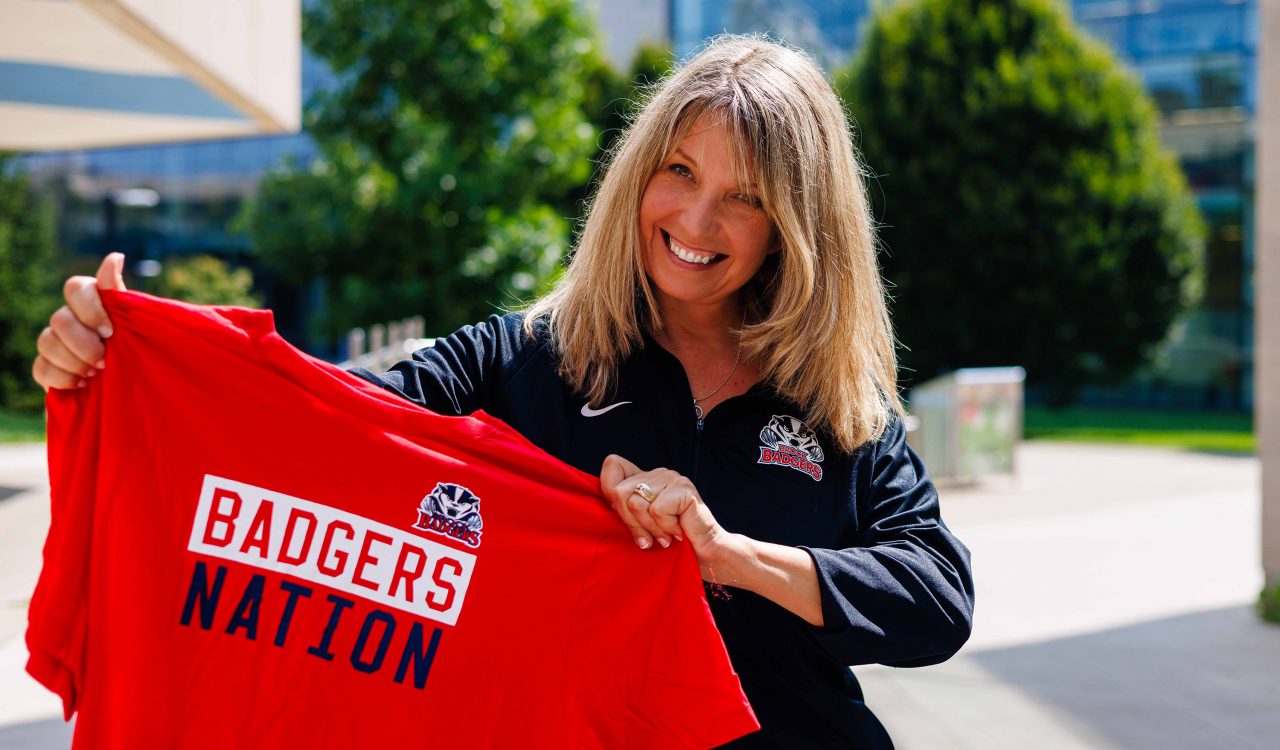 A smiling woman, wearing a Brock Badgers jacket, proudly holds up a red T-shirt that reads 'Badgers Nation' in bold letters. The photo is taken outdoors on a sunny day with trees and modern buildings in the background.