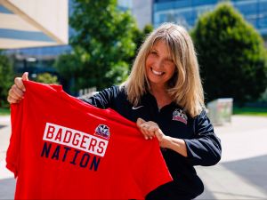 A smiling woman, wearing a Brock Badgers jacket, proudly holds up a red T-shirt that reads 'Badgers Nation' in bold letters. The photo is taken outdoors on a sunny day with trees and modern buildings in the background.