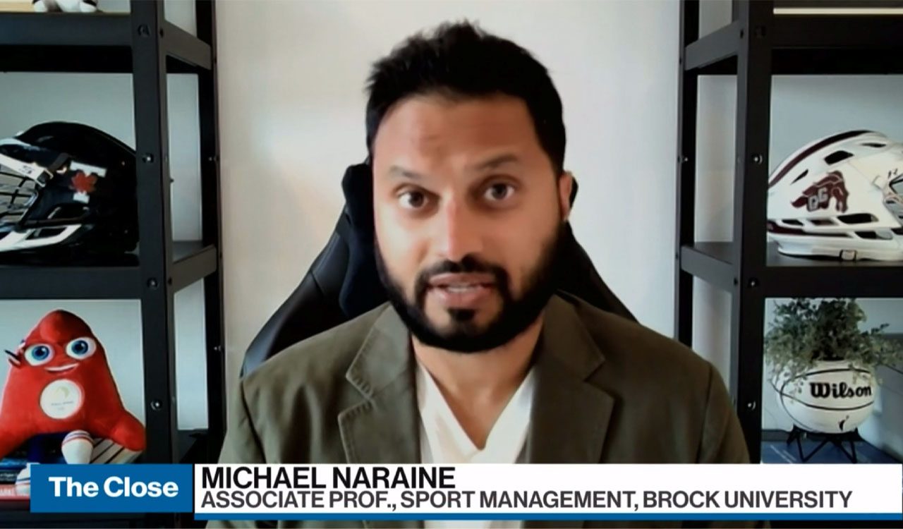 Michael Naraine conducts a virtual television interview flanked by two shelves with sports gear on them and wearing a beige blazer.