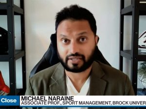 Michael Naraine conducts a virtual television interview flanked by two shelves with sports gear on them and wearing a beige blazer.