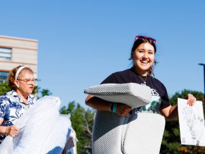 A woman carries a pillows with another woman following behind with arms full of luggage outside of Brock University’s Residence 8.