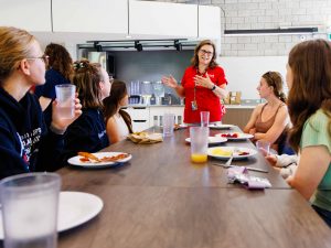 A woman stands in front of students speaking at the of a dining table inside Decew Dining Hall at Brock University.