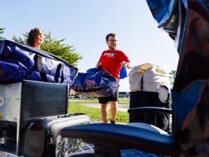 A man wearing a red Brock University shirt laughs outdoors while being surrounded by luggage, equipment and student living items.